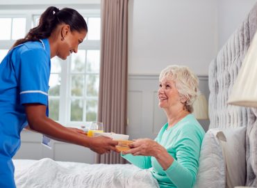 Female Care Worker In Uniform Bringing Senior Woman At Home Breakfast In Bed On Tray